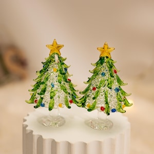 Op de afbeelding: Twee miniatuur kerstbomen van glas met heldere, getextureerde glazen takken en groene accenten. Elke boom is bekroond met een kleine gele glazen ster en versierd met kleine rode, blauwe en gele glazen bolletjes tussen de takken.