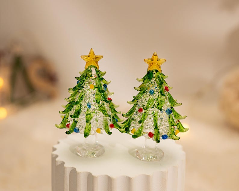 Op de afbeelding: Twee miniatuur kerstbomen van glas met heldere, getextureerde glazen takken en groene accenten. Elke boom is bekroond met een kleine gele glazen ster en versierd met kleine rode, blauwe en gele glazen bolletjes tussen de takken.