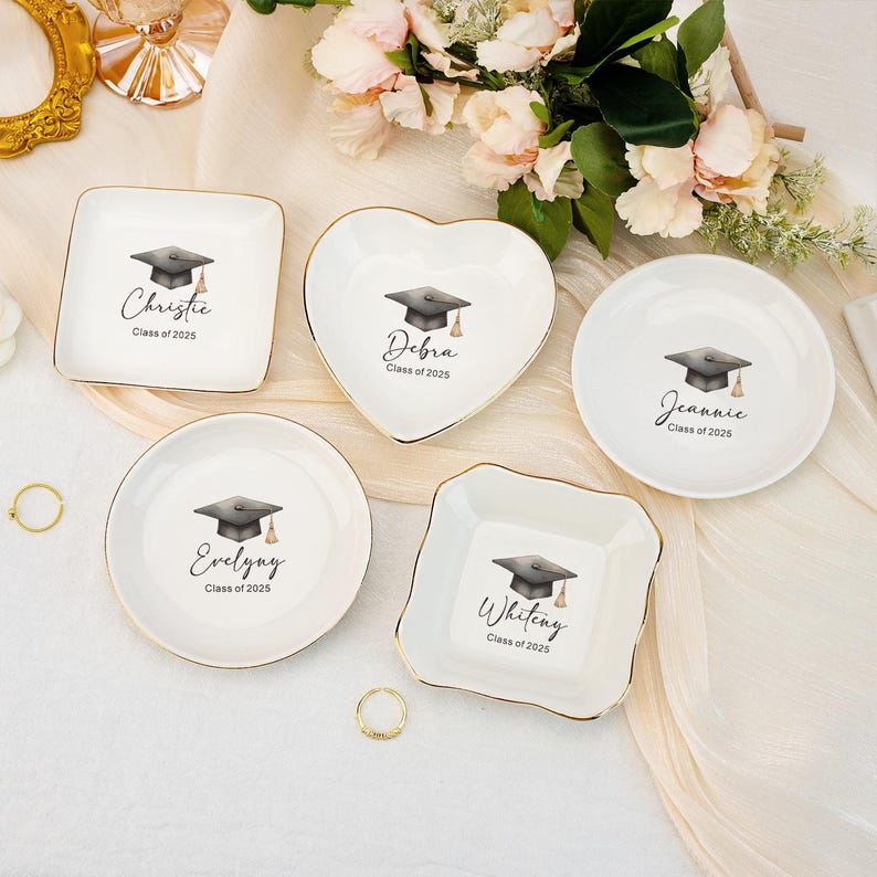 May include: Five white ceramic ring dishes with gold trim. Each dish features a black graduation cap and the name of a graduate, including Christie, Debra, Jeannie, Evelyny, and Whiteng, along with the year "Class of 2025".