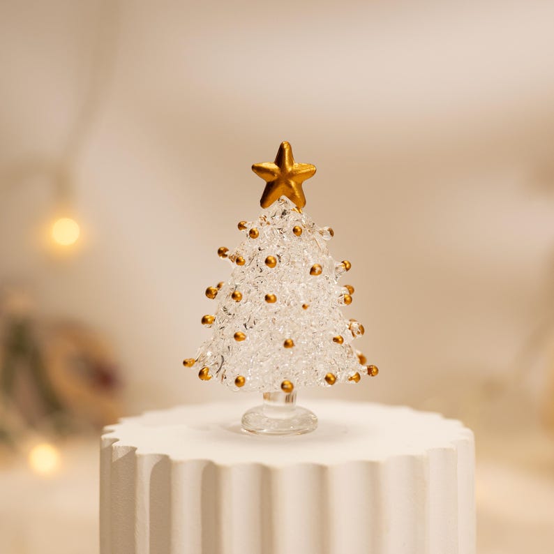 Op de afbeelding: Een kerstboomfiguur van helder glas met een getextureerd, kristallijn oppervlak. De boom is versierd met kleine, ronde goudkleurige kralen en wordt bekroond met een massieve goudkleurige ster. Het staat op een kleine, ronde glazen voet.