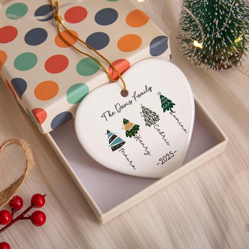 May include: A white heart-shaped ceramic ornament featuring the text The Davis Family and 2025. It displays four illustrated Christmas trees labeled with the names Maura, Henry, Cedric, and Lumina, suspended by a gold string.