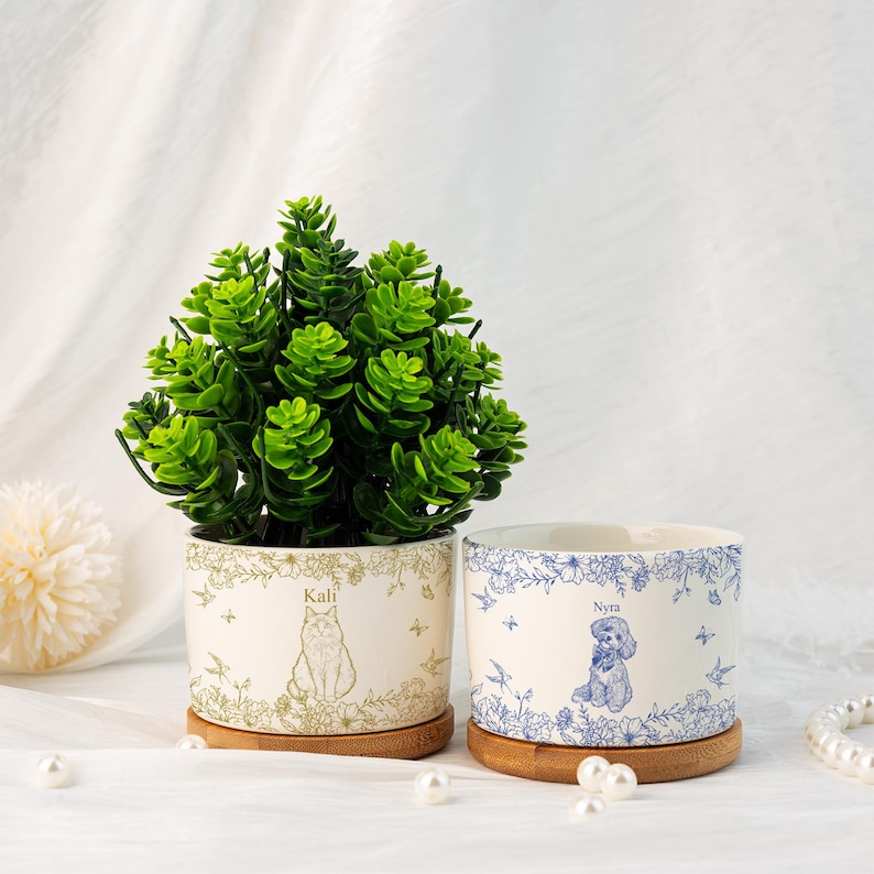 May include: Two ceramic plant pots with bamboo saucers. One pot features a cat illustration with the name "Kali", the other a dog illustration with the name "Nyra". Both pots have floral and bird designs. A green plant sits in one pot.