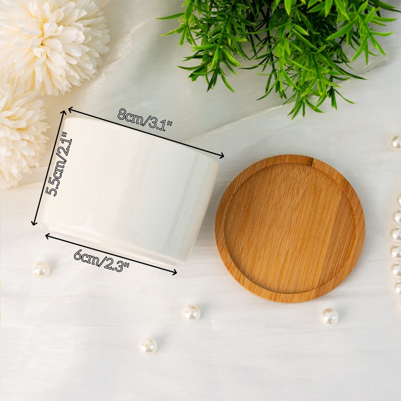 K&ouml;nnte beinhalten: Ein wei&szlig;er Keramik-Pflanztopf, 8 cm breit, 5,5 cm hoch und 6 cm tief, mit einer Untertasse aus Naturholz. Der Pflanztopf ist rechteckig mit abgerundeten Ecken. Die Untertasse ist rund mit einem erh&ouml;hten Rand. Dekorative Perlen und Gr&uuml;nzeug sind im Hintergrund.