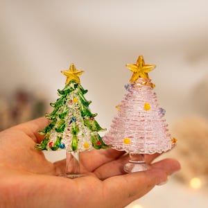 Op de afbeelding: Twee miniatuur kerstbomen van glas, &eacute;&eacute;n groen met veelkleurige glazen ornamenten en &eacute;&eacute;n lichtroze met kleine gele en blauwe accenten. Beide bomen hebben een doorzichtige glazen voet en een gele glazen ster als piek.
