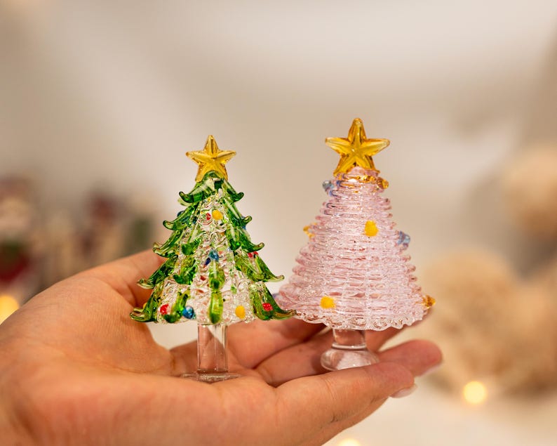 Op de afbeelding: Twee miniatuur kerstbomen van glas, &eacute;&eacute;n groen met veelkleurige glazen ornamenten en &eacute;&eacute;n lichtroze met kleine gele en blauwe accenten. Beide bomen hebben een doorzichtige glazen voet en een gele glazen ster als piek.