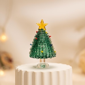 Op de afbeelding: Een miniatuur kerstboomfiguur van glas met een conisch groen lichaam met spiraalvormige glasstructuur, versierd met kleine veelkleurige glaskralen als ornamenten en bekroond met een gele glasster op een doorzichtige glazen voet.