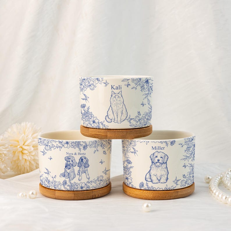 May include: Three ceramic pet planters with bamboo saucers. Each planter features a blue illustration of a cat or dog with the pet's name. The planters are decorated with blue floral and bird designs. The names on the planters are "Kali", "Nyra & Hertz", and "Miller".