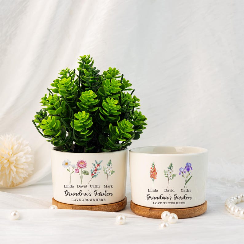 K&ouml;nnte beinhalten: Zwei wei&szlig;e Keramik-Blument&ouml;pfe mit floralen Designs und Bambus-Untersetzern. Die T&ouml;pfe tragen den Text "Grandma's Garden" und "LOVE GROWS HERE". Ein Topf enth&auml;lt eine gr&uuml;ne Pflanze. Namen sind &uuml;ber dem Text aufgedruckt.