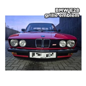 Peut inclure: Une voiture rouge BMW E28 avec le texte "BMW E28 grille emblem" en blanc. La voiture a des phares ronds, une calandre noire et un pare-chocs noir. La plaque d'immatriculation est partiellement masquée.