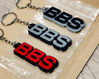 May include: Three keychains with the letters "BBS" in a layered design. Two keychains have gray letters with black outlines, and one has red letters with a black outline. Each keychain has a silver keyring and chain.