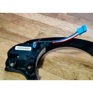 Puede incluir: Un módulo de control del volante de plástico negro con un conector de cable azul, rojo y blanco. El módulo tiene una etiqueta de código de barras con el número "70610700".