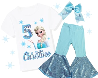 May include: A white t-shirt with a graphic of Elsa from Frozen, the number 5, and the name "Christine" in blue. The outfit includes light blue sequin pants and a matching bow with the name "Christine".