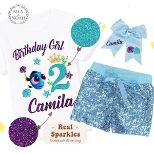 May include: A white t-shirt, blue sequin shorts, and a light blue bow set. The shirt features the text "Birthday Girl", the number "2", the name "Camila", and a cartoon fish. The shorts and bow are light blue.