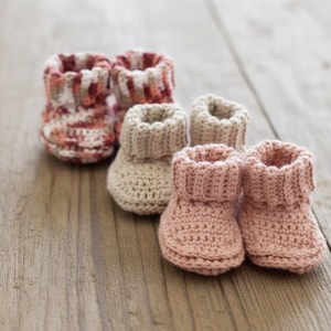 May include: A collection of six handmade baby booties in various colors. The booties are crocheted with a ribbed cuff and a rounded toe. Colors include a mix of red, white, and peach, as well as solid beige and pink. The booties are arranged on a wooden surface.