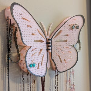 May include: A pink butterfly-shaped jewelry organizer with a dark gray outline. The wings have a perforated design and hold various necklaces and earrings. The jewelry organizer is mounted on a wall.