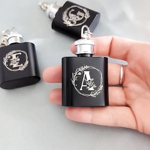 Puede incluir: Tres matraces de metal negro con tapas plateadas y diseños florales grabados. Los matraces tienen las letras "E", "G" y "A" en el centro de los diseños.