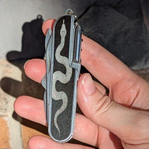 May include: A silver and black pocket knife with a detailed snake design. The snake is etched in white on a dark wood grain background. The knife has multiple tools and a keyring attachment.