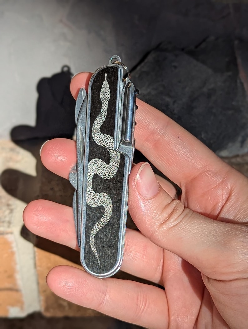 May include: A silver and black pocket knife with a detailed snake design. The snake is etched in white on a dark wood grain background. The knife has multiple tools and a keyring attachment.