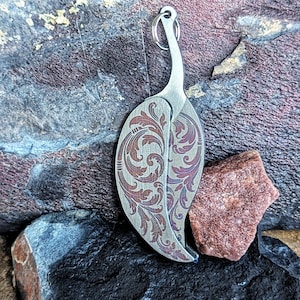 May include: A leaf-shaped pendant with a decorative floral pattern in shades of brown and purple. The pendant is made of metal and has a silver-coloured finish. It includes a small ring at the top for attaching to a chain or keychain.