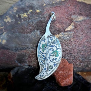 Op de afbeelding: Zilveren blad-vormige hanger met ingewikkelde bloemen- en wijnstokontwerpen in blauw en groen. De hanger heeft een lus aan de bovenkant voor een ketting. De achtergrond is een mix van donkerrode en grijze steen.