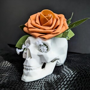 May include: A white skull-shaped planter with an orange rose and small white flowers. The skull has geometric facets and is set against a dark background. The rose is a vibrant orange, and the small flowers add a delicate touch.