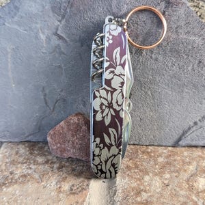 Puede incluir: Una navaja multiusos con diseño floral. La navaja tiene un fondo morado oscuro con patrones florales blancos. Incluye un sacacorchos, una hoja de cuchillo y otras herramientas. Un llavero de color cobre está adjunto.