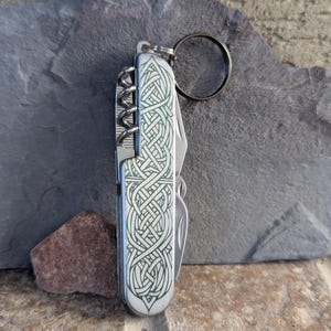 Puede incluir: Una multiherramienta plateada con un diseño de nudo celta verde. La herramienta incluye un sacacorchos, un cuchillo y otros implementos. Se adjunta un llavero negro. La herramienta está sobre una superficie de piedra gris.