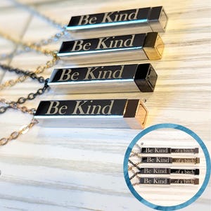 May include: Four silver necklaces with a rectangular pendant that says "Be Kind". Each pendant has a different colored accent on the end. The necklaces are on a light wood surface.