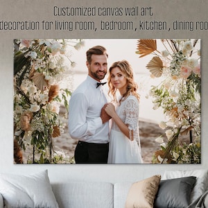 May include: A wedding photo of a couple standing in front of a floral archway. The bride is wearing a white lace dress and the groom is wearing a white shirt and black bow tie. The archway is decorated with white flowers, greenery, and dried palm leaves.