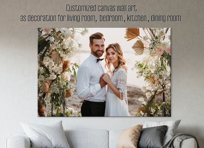 May include: A wedding photo of a couple standing in front of a floral archway. The bride is wearing a white lace dress and the groom is wearing a white shirt and black bow tie. The archway is decorated with white flowers, greenery, and dried palm leaves.