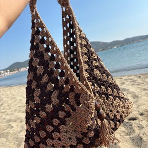 May include: A handmade crochet tote bag in shades of brown. The bag features a granny square pattern and long straps. Wooden beads and a tassel adorn the side. The bag is held up against a beach backdrop with a blue sky and ocean.