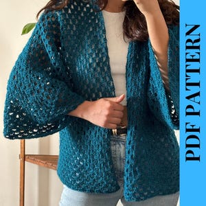 Peut inclure: Un cardigan en crochet bleu sarcelle avec un motif ajouré. Le cardigan a des manches larges et est ouvert sur le devant. Le texte "PDF PATTERN" est affiché verticalement sur un fond bleu. Le cardigan est porté sur un haut blanc et un jean bleu.