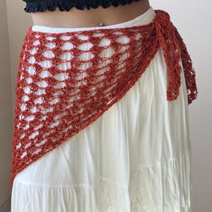 May include: A rust-colored, open-weave crochet wrap skirt featuring a diamond-patterned mesh design. The garment is tied at the side with a matching crochet cord and is layered over a solid white, tiered, floor-length skirt with a gathered hem.