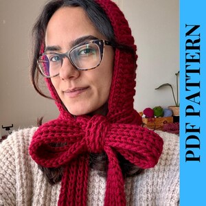 May include: A knitted red hood and scarf set with a large bow detail. The hood is pulled up, and the scarf is wrapped around the neck. The text "PDF PATTERN" is on a blue background.