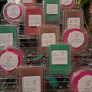May include: A selection of scented wax melts in clear packaging, with labels detailing scents such as "Self Love," "Apple Pie," and "Chai Spice." The melts are in various colours, including green, pink, red, and brown. The display is arranged on a tiered shelf.
