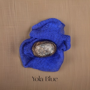 May include: A dark brown bar of soap resting in a blue mesh bag. The text "Yola Blue" is printed below the bag.
