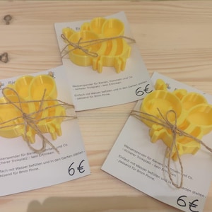 Può includere: Tre abbeveratoi da giardino gialli a forma di ape, legati con spago, ciascuno su una carta bianca con testo in tedesco. Gli abbeveratoi sono in plastica e sono progettati per le api. Su una superficie di legno chiaro.