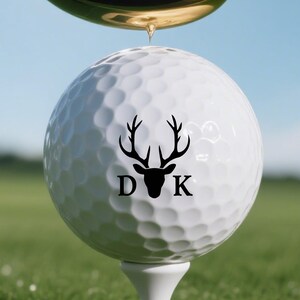 Könnte beinhalten: Ein weißer Golfball mit einem schwarzen Hirschkopf-Design und den Initialen "DK" balanciert auf einem Tee, mit einem Golfschläger darüber. Die Szene spielt sich auf einem grünen Golfplatz unter blauem Himmel ab.