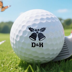 Könnte beinhalten: Ein weißer Golfball mit einem schwarzen Glocken- und Stechpalmen-Design und den Buchstaben "D&K" liegt auf grünem Gras neben einem Golfschläger. Schmetterlinge fliegen im Hintergrund unter blauem Himmel. Der Golfschläger hat einen roten Akzent.