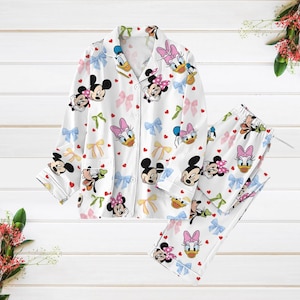 May include: White pyjamas with a button-up top and long trousers. The pyjamas feature a repeating pattern of Mickey Mouse, Minnie Mouse, Donald Duck, and Daisy Duck characters with pink and yellow bows.