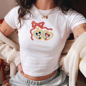 Op de afbeelding: Witte crop top met een Mickey en Minnie Mouse ontwerp. Het ontwerp toont Mickey en Minnie in hartvormige frames met een rode strik. De top heeft korte mouwen en is gemaakt van een zachte stof.