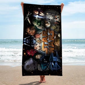 May include: A black rectangular beach towel with a Star Wars collage design. The towel features various characters and the Star Wars logo in gold and orange. The towel is held up against a beach and ocean backdrop.