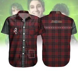 May include: A short-sleeved button-up shirt with a red and black plaid pattern. The shirt features black accents on the sleeves and collar, with decorative elements including a gear and a small rectangular patch. The shirt has a brown leather-like patch on the back.