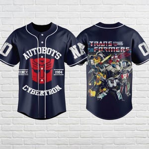 May include: Navy blue baseball jersey featuring the Autobots logo and text "AUTOBOTS CYBERTRON SINCE 1984". The back of the jersey displays a graphic of Transformers characters and the text "TRANSFORMERS". White trim and accents.