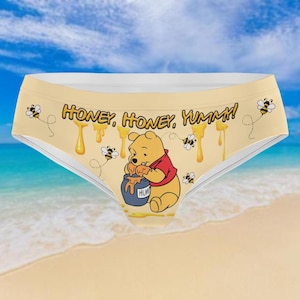May include: Light yellow underwear with a Winnie the Pooh design. The text "HONEY, HONEY, YUMMM!" is printed across the front, with bees and honey drips. The background is a beach scene. The character is wearing a red shirt and holding a blue honey pot.
