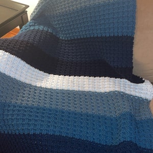 May include: A blue and white crocheted throw blanket with a textured pattern. The blanket is folded and draped over a sofa.