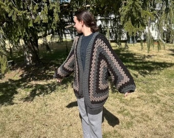 Könnte beinhalten: Ein handgefertigter, offener Häkelcardigan in Grau-, Schwarz- und Beigetönen. Der übergroße Pullover hat ein geometrisches Muster und lange Ärmel. Die Person trägt eine graue Jogginghose und schwarze Schuhe.