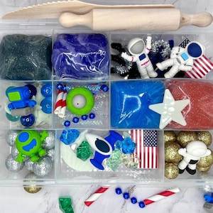 Puede incluir: Un contenedor de almacenamiento de plástico transparente con compartimentos llenos de varios artículos para una actividad de juego sensorial con temática espacial. El contenedor incluye plastilina azul, roja y negra, figuras de astronautas, un rodillo y otros juguetes pequeños.
