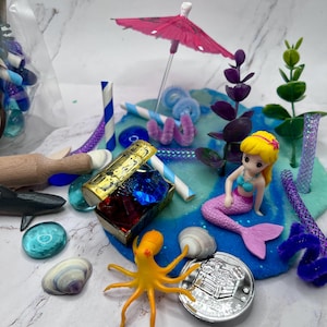 May include: A miniature mermaid scene with a pink parasol, a treasure chest, and an octopus. The scene includes a mermaid figurine, blue and green playdough, and various decorative elements like seashells and glass gems.
