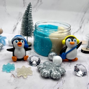 May include: A winter-themed sensory kit featuring two penguin figurines, a jar of blue and beige sensory dough, and decorative elements like snowflakes and miniature trees. The penguins wear hats and headphones. The kit includes a rolling pin.